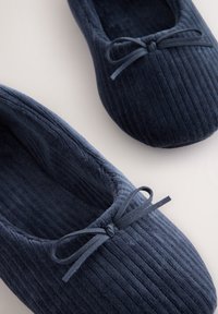 Paire de chaussons en velours côtelé bleu marine avec petits nœuds en tissu sur le dessus, vus de dessus sur une surface claire.