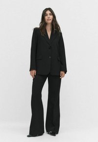 Mustat räätälöidyt bleiserit, joissa on yksi nappisulku, leikattu kaulus ja sivutaskut, yhdistettynä leveälahkeisiin mustiin housuihin ja teräviin mustiin kenkiin.