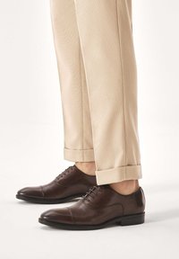 Rjavi usnjeni elegantni čevlji z gladko teksturo, okroglo konico, z vezalkami in subtilnimi šivi, kombinirani z zavihanimi bež hlačami.