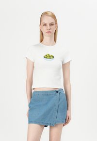Camiseta blanca de algodón de manga corta con un gráfico de un cuenco de aceitunas verdes. Combinada con una falda envolvente de mezclilla azul.