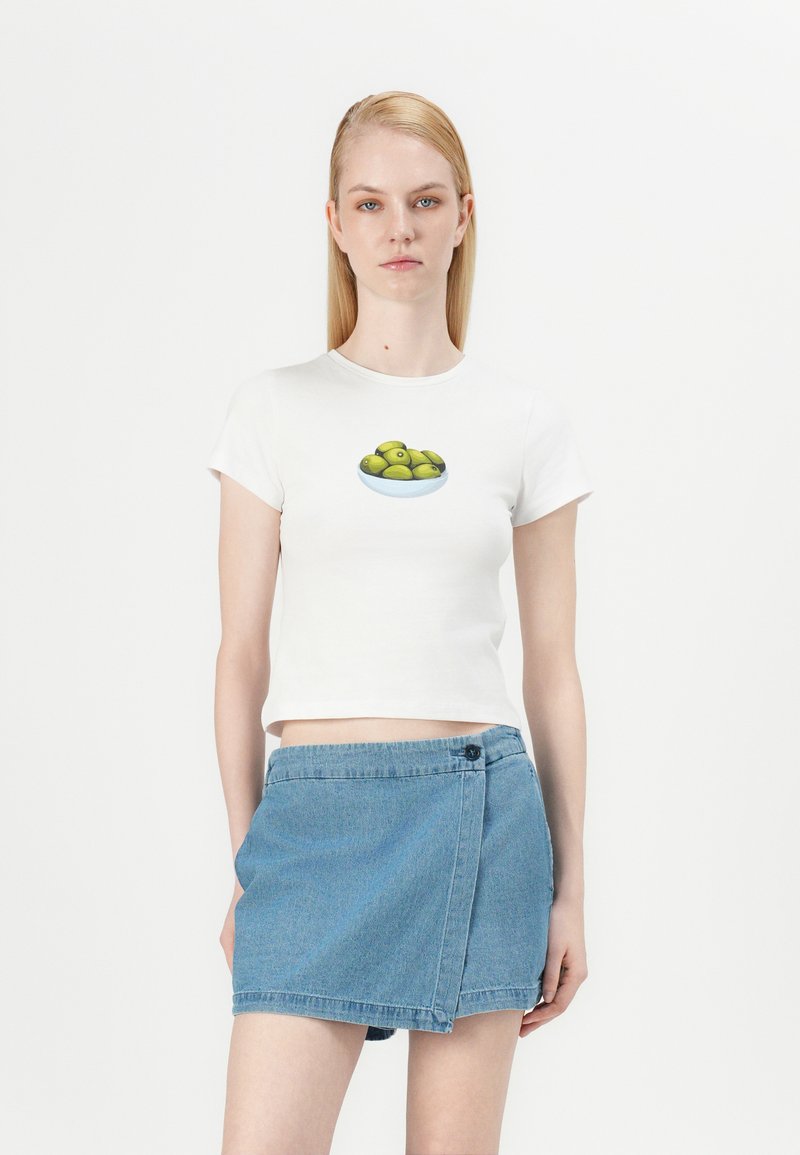 Camiseta blanca de algodón de manga corta con un gráfico de un cuenco de aceitunas verdes. Combinada con una falda envolvente de mezclilla azul.