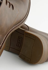 Zapatos de cuero marrón con textura suave, cordones y costuras blancas en contraste, con suelas de goma y una plantilla con marca.