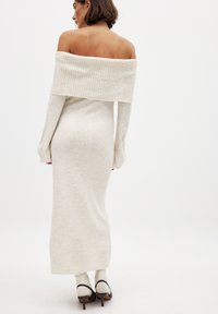 Abito in maglia off-the-shoulder color beige chiaro, con maniche lunghe, silhouette aderente, colletto e polsini a coste, abbinato a scarpe con tacco beige.