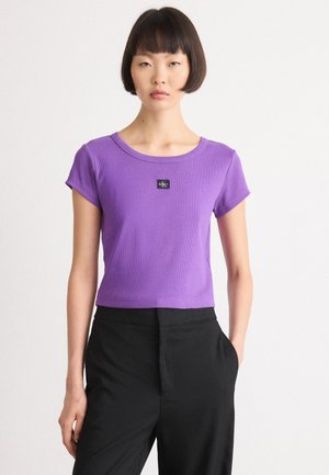 Purple ribbed short-sleeve T-shirt with a round neckline, featuring a black logo patch at the center. Paired with black trousers.