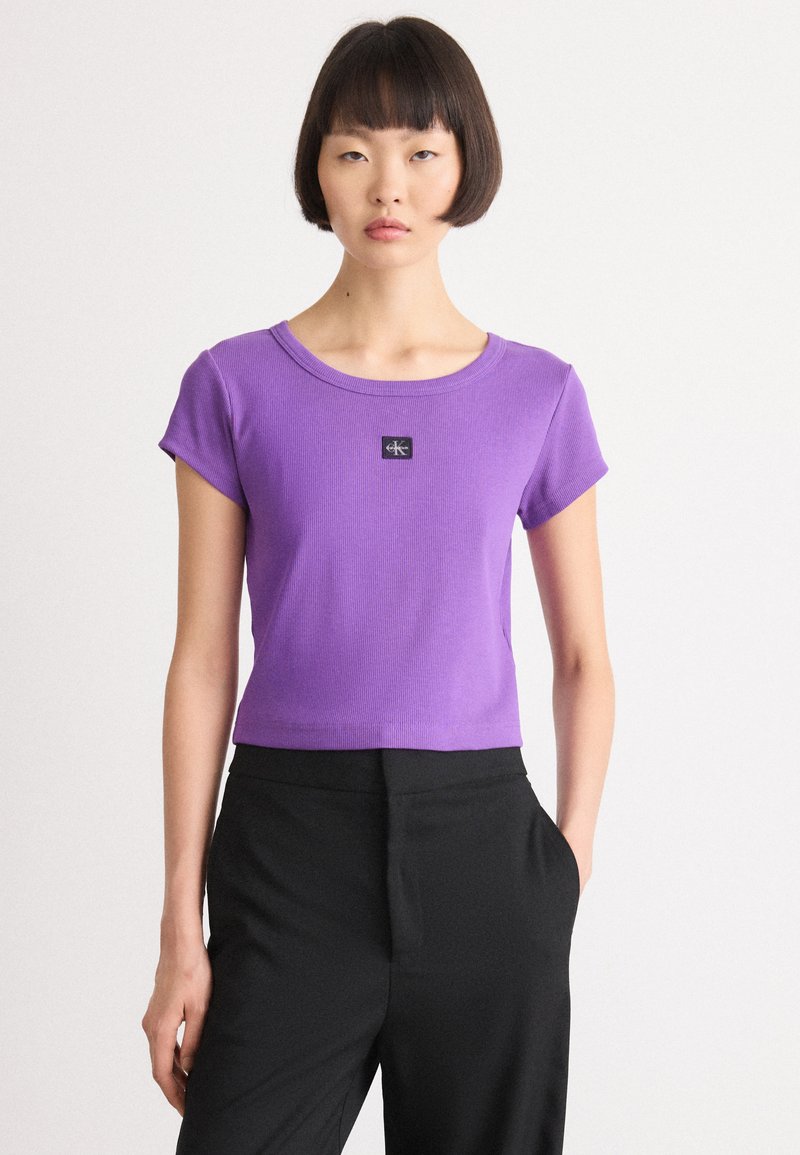 Purple ribbed short-sleeve T-shirt with a round neckline, featuring a black logo patch at the center. Paired with black trousers.