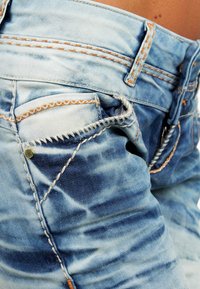 Jeans in denim azzurro chiaro con sezioni sbiadite, cuciture arancioni, una piccola tasca anteriore e dettagli in rivetto color oro.