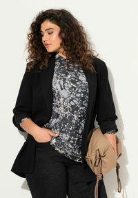 Sort blazer over en gråmønstret top, parret med sorte strukturerede bukser. Accessoriseret med en lysebrun toilettaske.