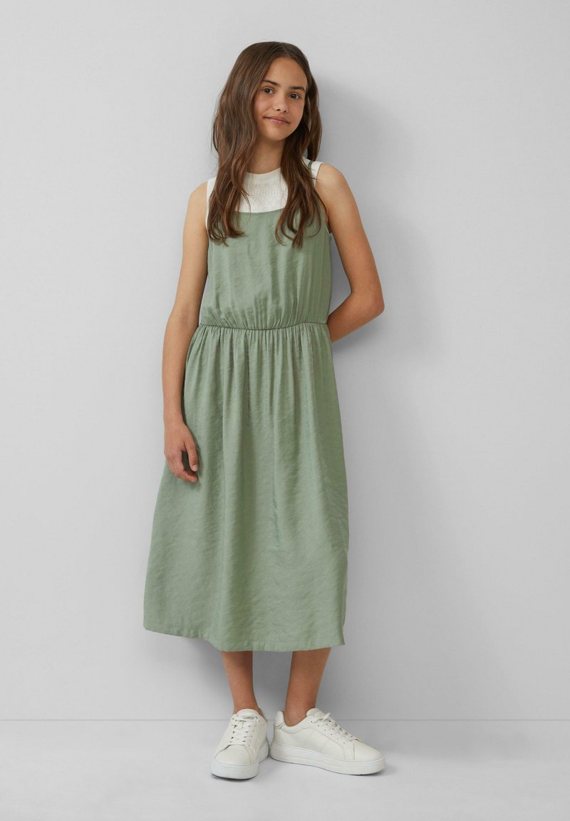 Grünes Midikleid aus leichtem Stoff, mit einem elastischen Taillengummi und einem entspannten Schnitt, kombiniert mit weißen Sneakers.