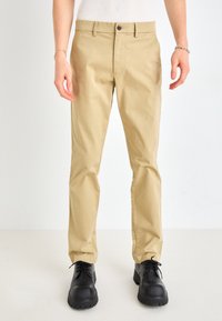 Beige bomullsbukser har en rett leggedesign, knappeåpning og frontlommer. Sammen med svarte sko er antrekket uformelt og allsidig.