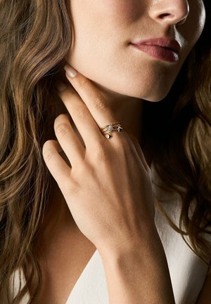 Frau mit langen braunen Haaren, die sanft ihren Hals berührt und drei goldene Ringe mit kleinen hängenden Anhängern an ihrem Finger trägt.