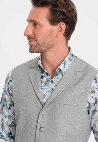 Gray textured vest with two buttons and a front pocket, worn over a patterned light blue floral shirt.