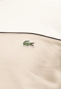 Κοντινό πλάνο μπεζ και εκρού υφάσματος με καφέ περιθώριο και ένα μικρό κεντημένο πράσινο λογότυπο κροκόδειλου.