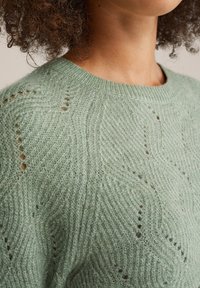 Maglione lavorato a maglia verde chiaro con un motivo ondulato e testurizzato e piccoli fori decorativi. Include un classico scollo a girocollo e polsini a coste.
