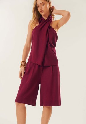 Femme portant un haut sans manches bordeaux à col halter et un short large assorti jusqu'aux genoux, accessoirisée de bracelets et boucles d'oreilles en or.