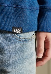 Sudadera azul texturizada con puños acanalados, con una etiqueta negra de Superdry. Combinada con jeans de mezclilla azul claro, mostrando detalle de bolsillo.