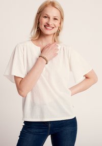 Blusa bianca di lino con maniche corte e larghe e un collo rotondo. Il tessuto ha una sottile texture. Indossata con jeans blu scuri.