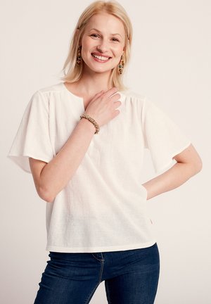 Blusa bianca di lino con maniche corte e larghe e un collo rotondo. Il tessuto ha una sottile texture. Indossata con jeans blu scuri.