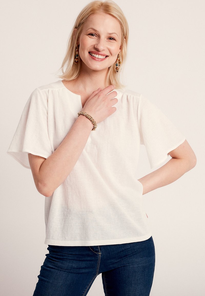 Blusa bianca di lino con maniche corte e larghe e un collo rotondo. Il tessuto ha una sottile texture. Indossata con jeans blu scuri.