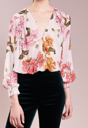 Persoon die een witte blouse draagt met roze en oranje bloemenpatronen en zwarte fluwelen broek, staand tegen een effen lichte achtergrond.