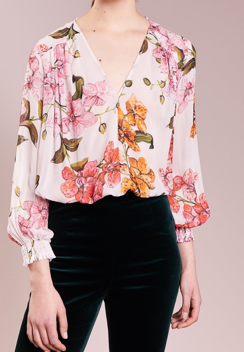 Personne portant un chemisier blanc avec des motifs floraux roses et oranges et un pantalon en velours noir, debout devant un fond clair uni.