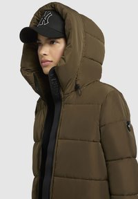 Mørkeoliven pufferjakke med stor hætte, med kviltet design, frontlynlås og logodetaljer. Akkompagneret af en sort kasket.