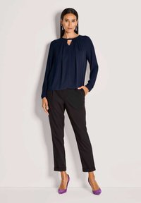 Marineblaues Langarmbluse mit Schlüsselloch-Ausschnitt, kombiniert mit schwarzen tapered Hosen und lila High Heels. Glattes Gewebe mit dezenten Drapierungselementen.