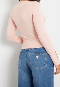 Pitkähihainen, vartalonmyötäinen yläosa pehmeästä vaaleanpunaisesta neuloskankaasta, yhdistetty sinisiin leveälahkeisiin farkkuihin, joissa kolmion muotoinen logo takataskussa.