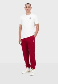 Camiseta blanca de algodón con un pequeño logo azul, combinada con pantalones de chándal rojos que presentan el mismo logo. Zapatillas blancas con detalles en dorado.