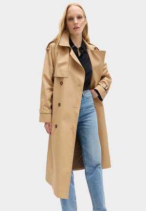 Kvinde iført en beige lang trenchcoat over en sort skjorte og blå jeans, stående mod en ensartet lys baggrund.