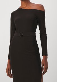 Černé šaty s dlouhým rukávem a off-shoulder designem, páskem v oblasti pasu s kulatou sponou, hladkou texturou a rozparkem na jedné straně.