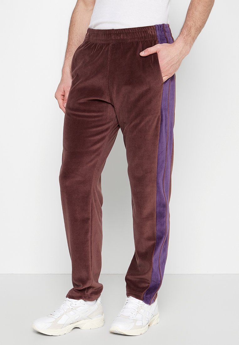 Pantalons de survêtement en velours bordeaux avec des rayures latérales violettes, dotés d'une taille élastique et de poches latérales. Texture lisse, coupe fuselée.