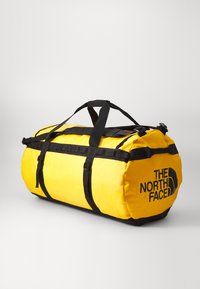 Bolsa de viaje amarilla hecha de material duradero, con correas y hebillas negras, forma cilíndrica y un logotipo en letras negras en negrita.