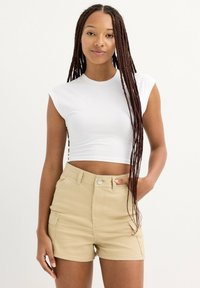 Top corto sin mangas blanco combinado con pantalones cortos beige de cintura alta que cuentan con bolsillos laterales, costuras en contraste y una textura de tela suave.