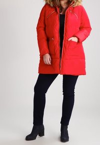 Červená prošívaná bunda s kapucí lemovanou umělou kožešinou, zipovými kapsami a lesklým zipem. Nositelná přes černé legíny a kotníkové boty.