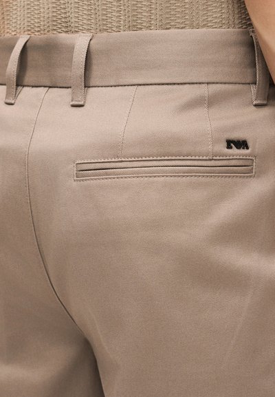 Κοντινό πλάνο τσέπης μπεζ παντελονιού με θηλιές για ζώνη και μαύρο λογότυπο "EVA" στην πίσω δεξιά πλευρά.