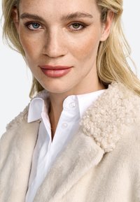 Manteau beige avec un col texturé et duveteux sur une chemise blanche à boutons, présentant un tissu doux et une coupe ajustée. Un maquillage naturel met en valeur les traits.