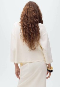 Haut blanc surdimensionné avec des manches trois-quarts, associé à une jupe assortie. Texture remarquable provenant de cheveux bouclés tombant le long du dos.