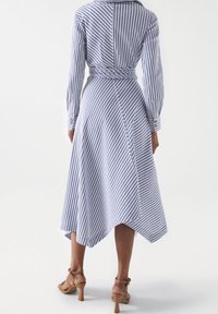 Vestido de manga larga a rayas en azul claro y blanco, con cintura cruzada y bajo asimétrico. Detallado con puños con botones, confeccionado en tela ligera.