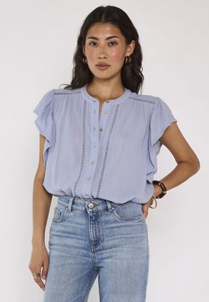 Femme aux longs cheveux foncés porte un chemisier bleu clair à volants et un jean bleu, debout avec une main dans la poche sur un fond uni.