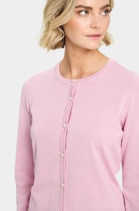 Cardigan rose clair en tissu doux, avec un col rond et des boutons ressemblant à des perles sur le devant. Conçu avec des manches longues.