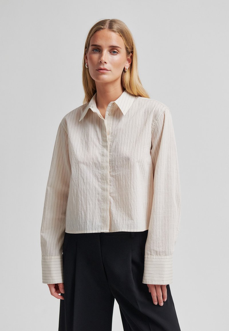 Jeune femme aux cheveux blonds longs portant une chemise ample à fines rayures beige clair boutonnée et un pantalon noir à jambes larges sur un fond uni.