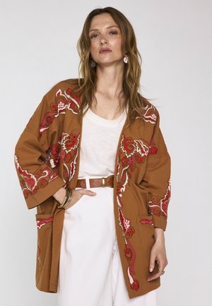 Frau trägt weißen Oberteil und Hose mit braun bestickter Kimono-Jacke, ergänzt durch Ohrringe, Armreifen und Gürtel, posiert vor einfachem Hintergrund.
