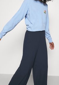 Personne portant une chemise à manches longues bleu clair avec un petit motif et un pantalon large taille haute bleu marine sur fond uni.