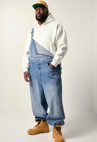Mand med gul cap, hvid hættetrøje, løse denim overalls med én strop og brune arbejdssko står mod en ensfarvet lys baggrund.