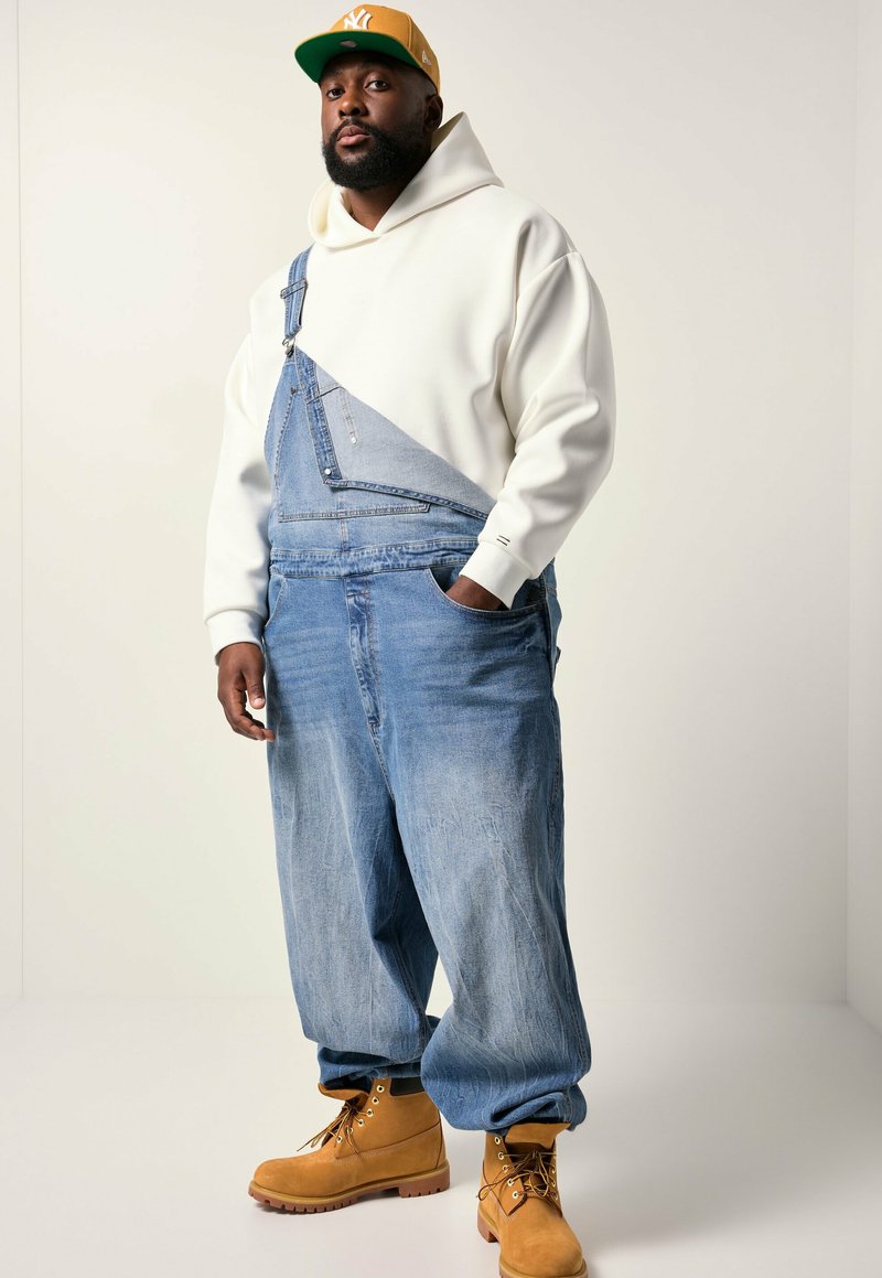 Mand med gul cap, hvid hættetrøje, løse denim overalls med én strop og brune arbejdssko står mod en ensfarvet lys baggrund.