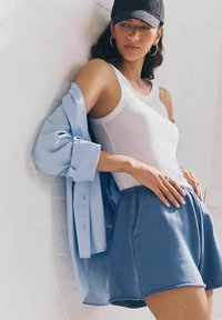 Canottiera bianca a coste sotto una camicia oversize azzurro chiaro, abbinata a pantaloni corti in cotone blu con cordoncino e tasche.