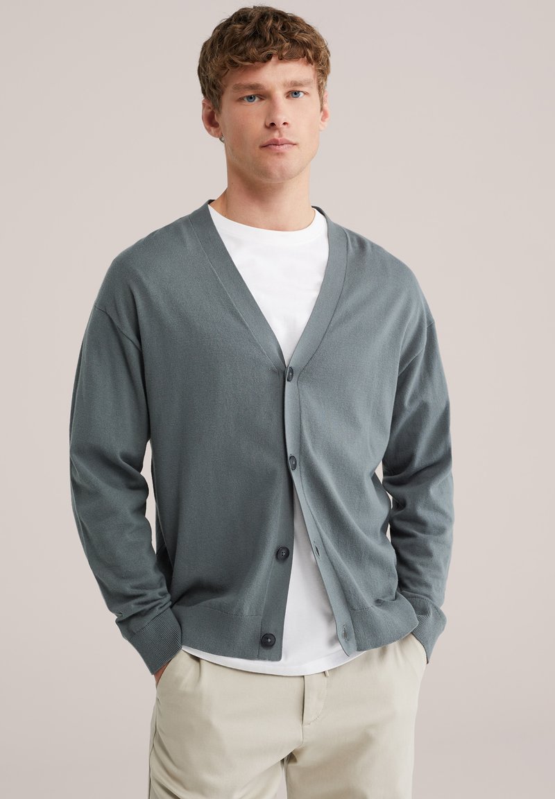 Jeune homme portant un cardigan gris boutonné sur un T-shirt blanc et un pantalon beige, debout les mains dans les poches devant un fond neutre.