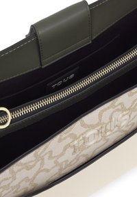 Primer plano de un bolso beige y oliva abierto que muestra un cierre dorado, un interior negro y el logo grabado "TOUS" en un panel exterior con patrón.