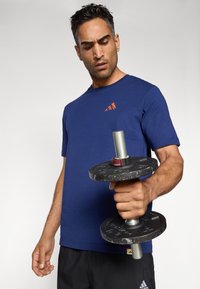 Man in een blauw shirt en zwarte shorts houdt een zwart metalen halter vast en kijkt ernaar tegen een eenvoudige lichte achtergrond.