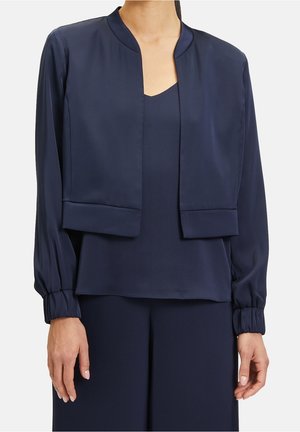 Mujer con una chaqueta corta de manga larga azul marino sobre un top sin mangas a juego y pantalones de pierna ancha, con puños recogidos en la chaqueta.